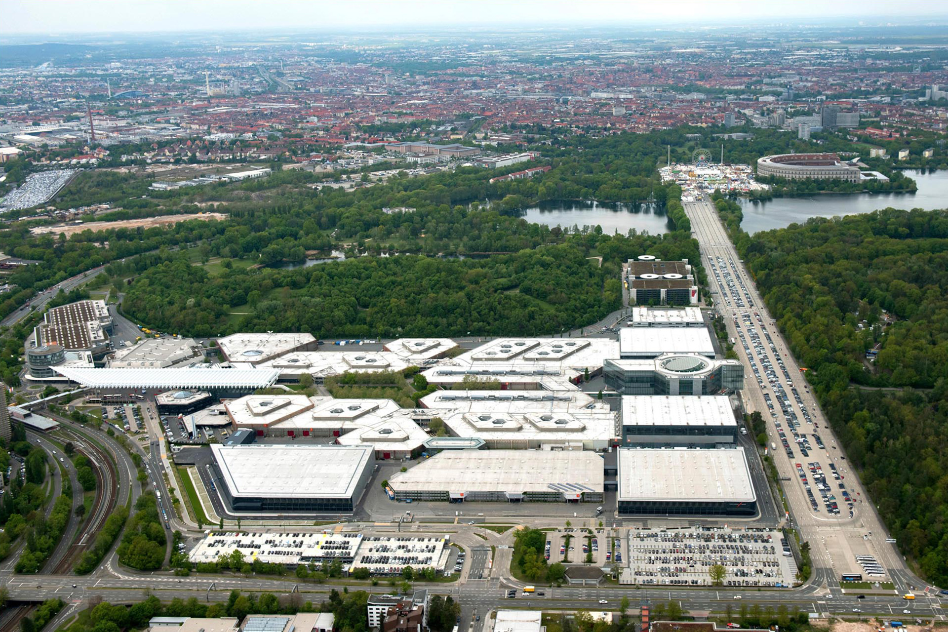Luftaufnahme des Messe Nürnberg mit seinen diversen Hallen. Rechts erkennt man einen großen Parkplatz, dahinter ein kleines Waldstück bevor die Stadt zu sehen ist.